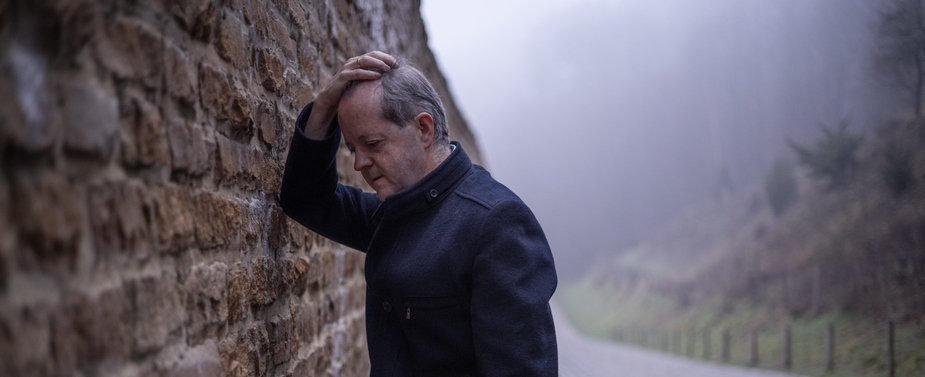 While leaning against a worn out wall, a senior man alone in misty forest, lonely and abandoned in gloomy atmospheric mood